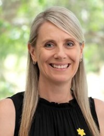 Smiling person with blonde hair, wearing a black sleeveless top and a yellow flower pin, outdoors.