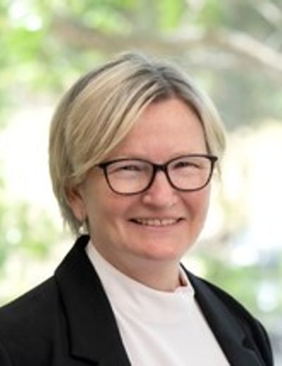 Smiling person with short blonde hair and glasses, wearing a black blazer and white top, outdoors.