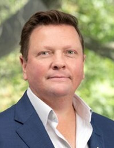 Portrait of a person in a blue blazer and white shirt, outdoors against green foliage.