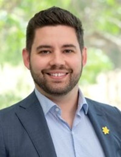 Smiling person in a blue blazer and light shirt, with a lapel pin, outdoors.