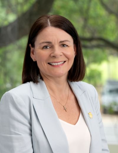 Smiling person with shoulder-length dark hair, wearing a navy blazer and pearl necklace.