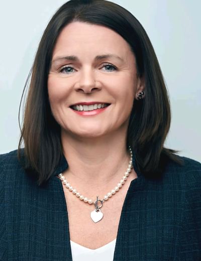 Smiling person with shoulder-length dark hair, wearing a navy blazer and pearl necklace.