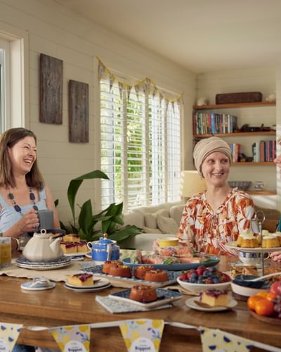 Four people share tea and cakes around a wooden table in a sunlit, plant-filled dining room.