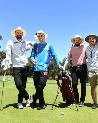 Four men standing and smiling, wearing sun protective clothing, at a golf course with golf clubs.
