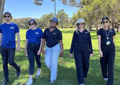 Five people walk through a sunny park in blue tops and hats under trees.