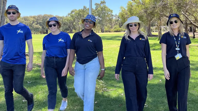Five people walk through a sunny park in blue tops and hats under trees.