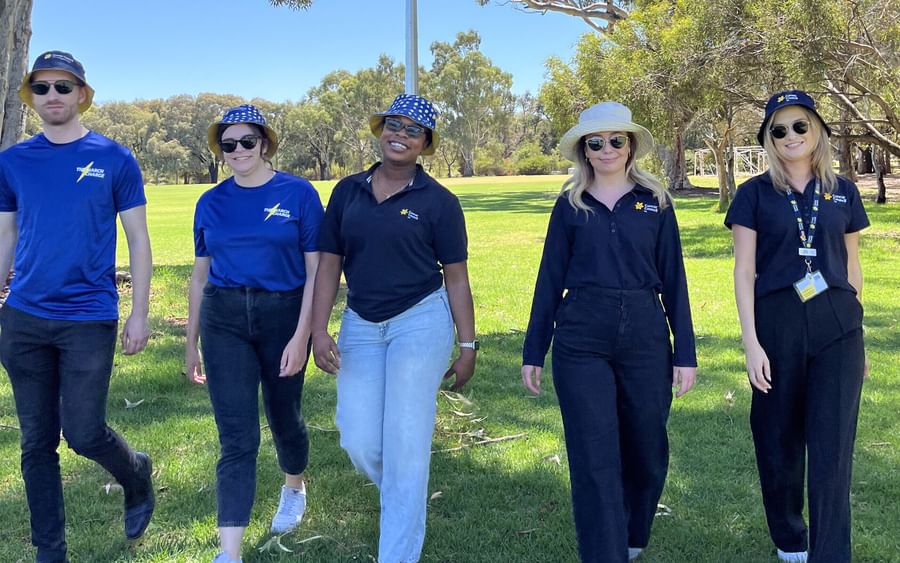 Five people walk through a sunny park in blue tops and hats under trees.
