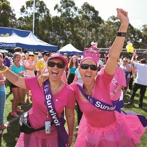 Two participants in pink outfits with sashes cheer at a Cancer Council event in a park.
