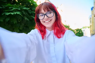 Person with red hair and glasses taking a sunny outdoor selfie, smiling in a white shirt.