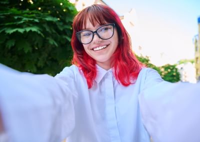 Person with red hair and glasses taking a sunny outdoor selfie, smiling in a white shirt.