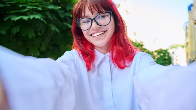 Person with red hair and glasses taking a sunny outdoor selfie, smiling in a white shirt.
