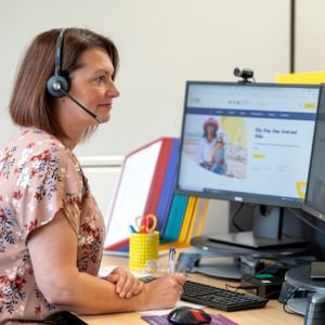 Person with short brown hair in a floral blouse wearing a headset, at a desk with two monitors.