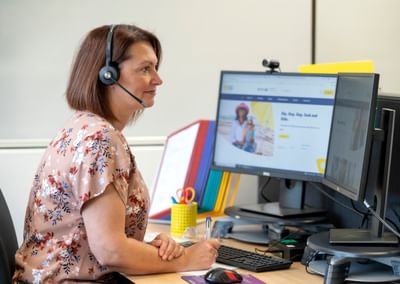 Person with short brown hair in a floral blouse wearing a headset, at a desk with two monitors.