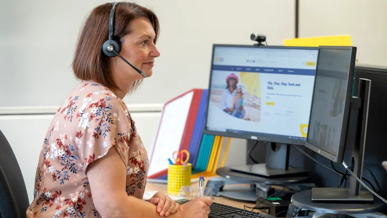 Person with short brown hair in a floral blouse wearing a headset, at a desk with two monitors.