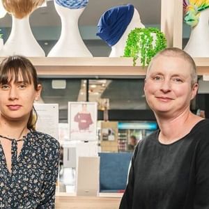 Two people sit together in a bright shop with a wig display behind them, smiling.