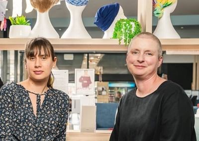 Two people sit together in a bright shop with a wig display behind them, smiling.