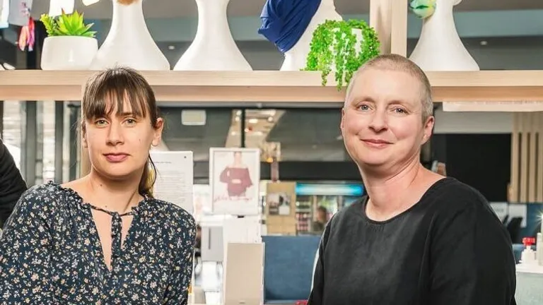 Two people sit together in a bright shop with a wig display behind them, smiling.