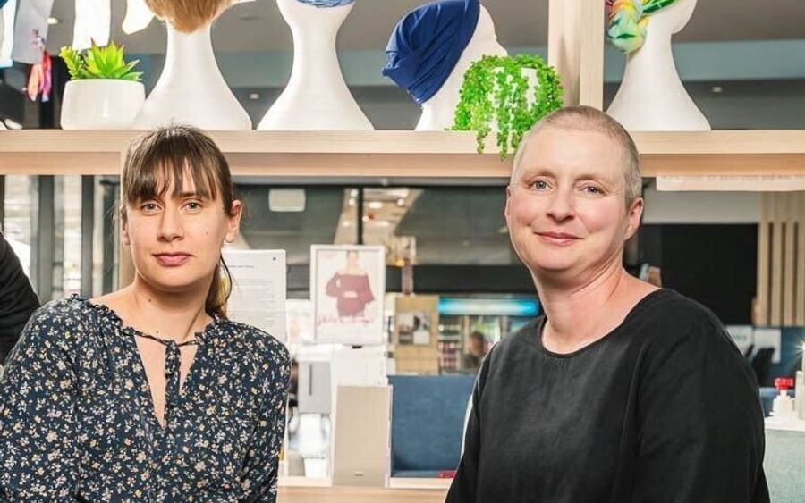 Two people sit together in a bright shop with a wig display behind them, smiling.