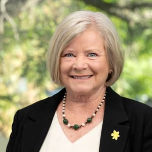 Smiling person with short grey hair in a black blazer and green beaded necklace; outdoor background.