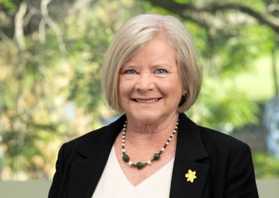 Smiling person with short grey hair in a black blazer and green beaded necklace; outdoor background.