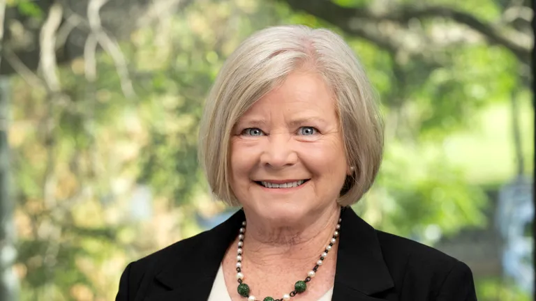 Smiling person with short grey hair in a black blazer and green beaded necklace; outdoor background.