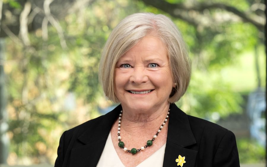 Smiling person with short grey hair in a black blazer and green beaded necklace; outdoor background.