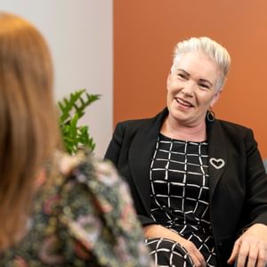 Smiling person with white hair in a black blazer and checkered dress chats with another person.