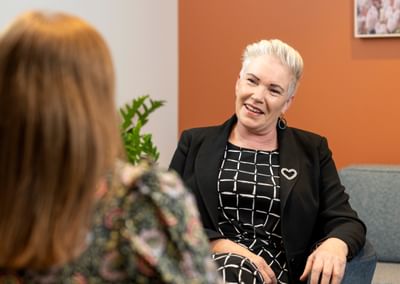 Smiling person with white hair in a black blazer and checkered dress chats with another person.