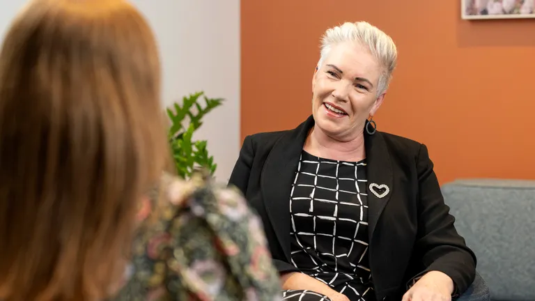 Smiling person with white hair in a black blazer and checkered dress chats with another person.