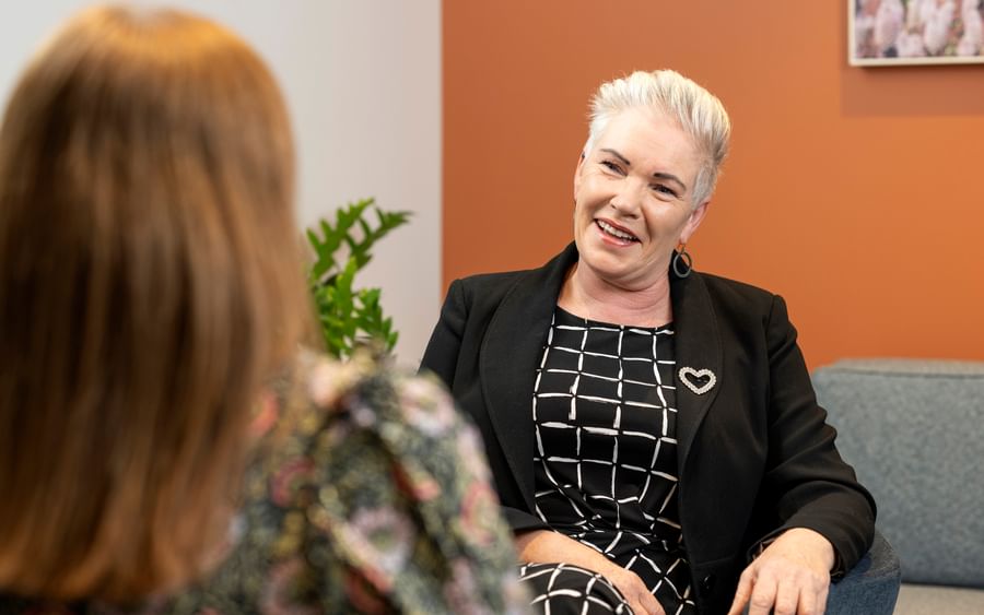 Smiling person with white hair in a black blazer and checkered dress chats with another person.