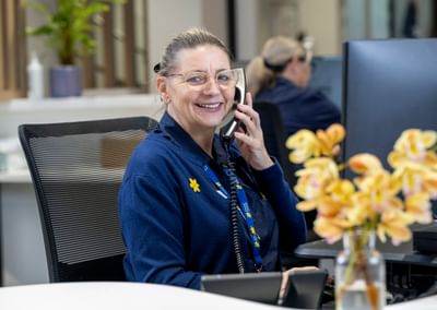 Smiling staff member with glasses on a phone at a reception desk in an office, a vase of flowers.
