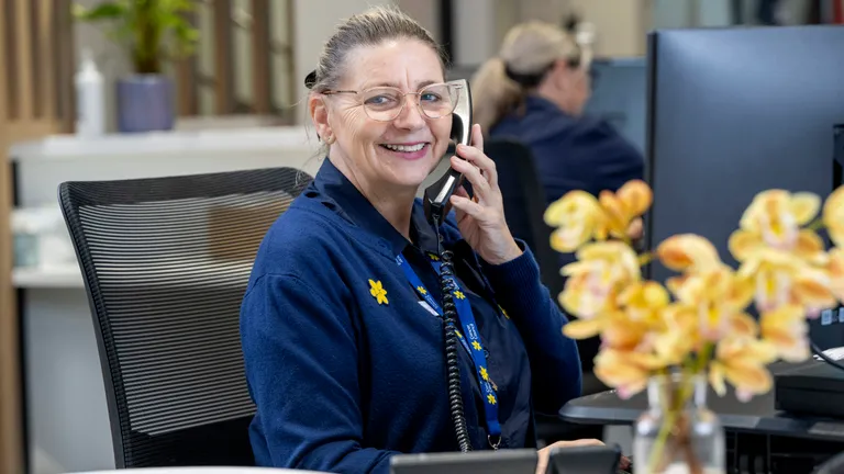 Smiling staff member with glasses on a phone at a reception desk in an office, a vase of flowers.