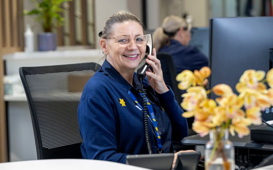 Smiling staff member with glasses on a phone at a reception desk in an office, a vase of flowers.