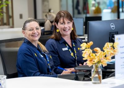 Two staff members in blue uniforms sit at a reception desk, smiling, with a vase of yellow flowers.