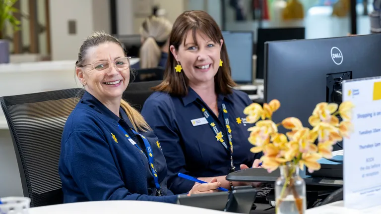 Two staff members in blue uniforms sit at a reception desk, smiling, with a vase of yellow flowers.