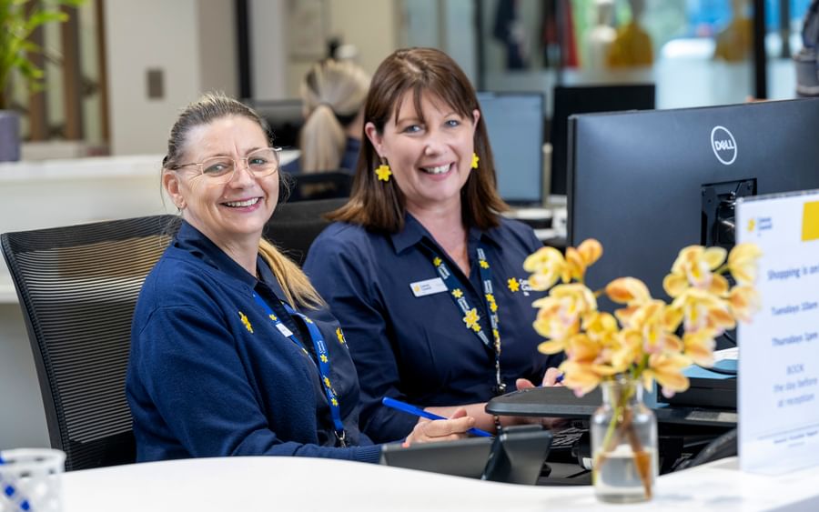 Two staff members in blue uniforms sit at a reception desk, smiling, with a vase of yellow flowers.