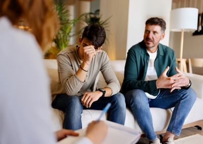 Two on a sofa; one distressed with a hand over their face, while another person takes notes.