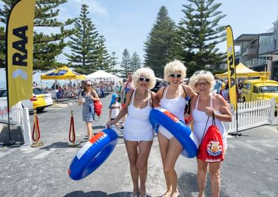 Three people in white outfits and blonde wigs pose with blue inflatable rings at a street festival.