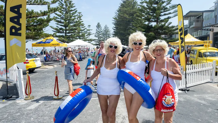 Three people in white outfits and blonde wigs pose with blue inflatable rings at a street festival.