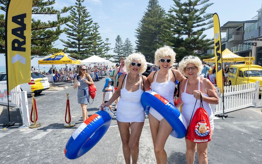 Three people in white outfits and blonde wigs pose with blue inflatable rings at a street festival.
