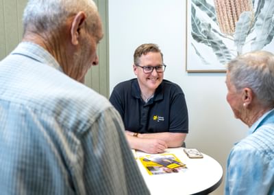 Person in a black Cancer Council polo smiles while chatting with two older adults at a round table with brochures.
