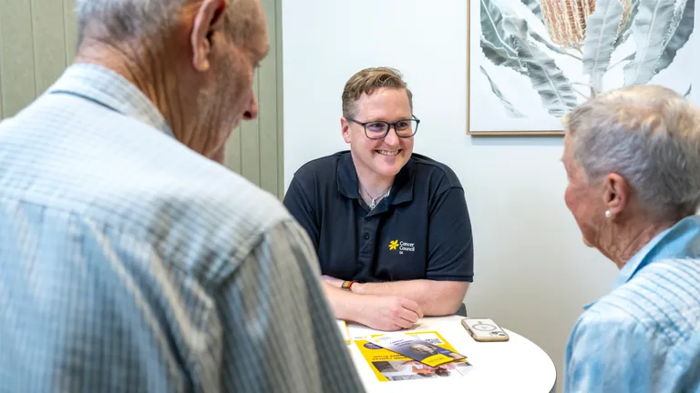 Person in a black Cancer Council polo smiles while chatting with two older adults at a round table with brochures.