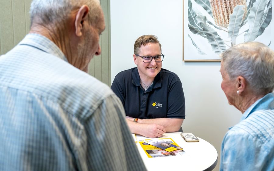 Person in a black Cancer Council polo smiles while chatting with two older adults at a round table with brochures.