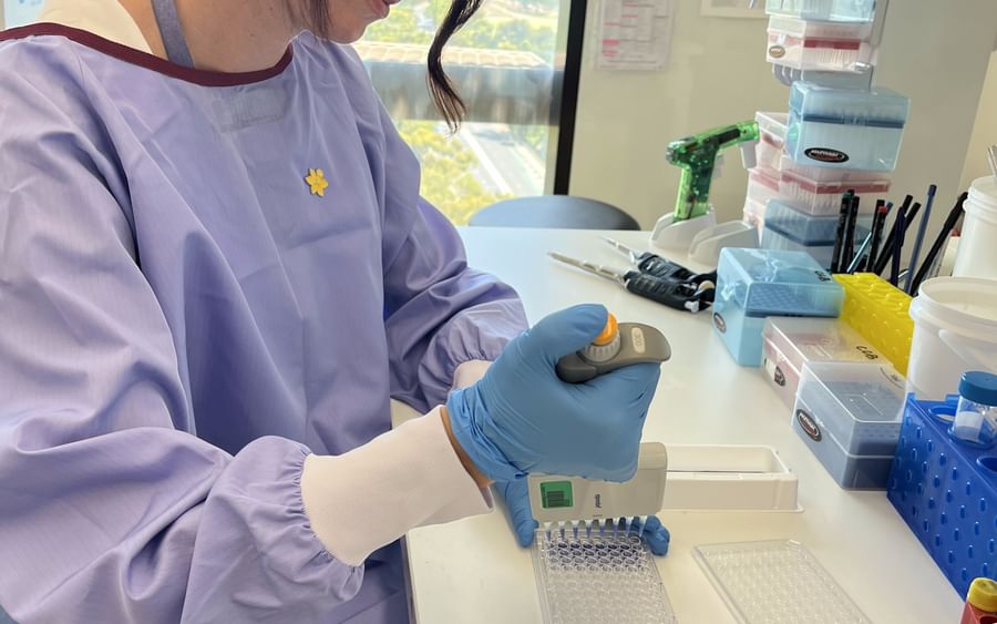 Lab researcher in a purple gown and blue gloves pipetting into a 96-well plate.