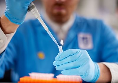 Person in blue lab coat and gloves uses a pipette to transfer liquid into a small tube.