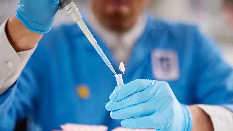 Person in blue lab coat and gloves uses a pipette to transfer liquid into a small tube.