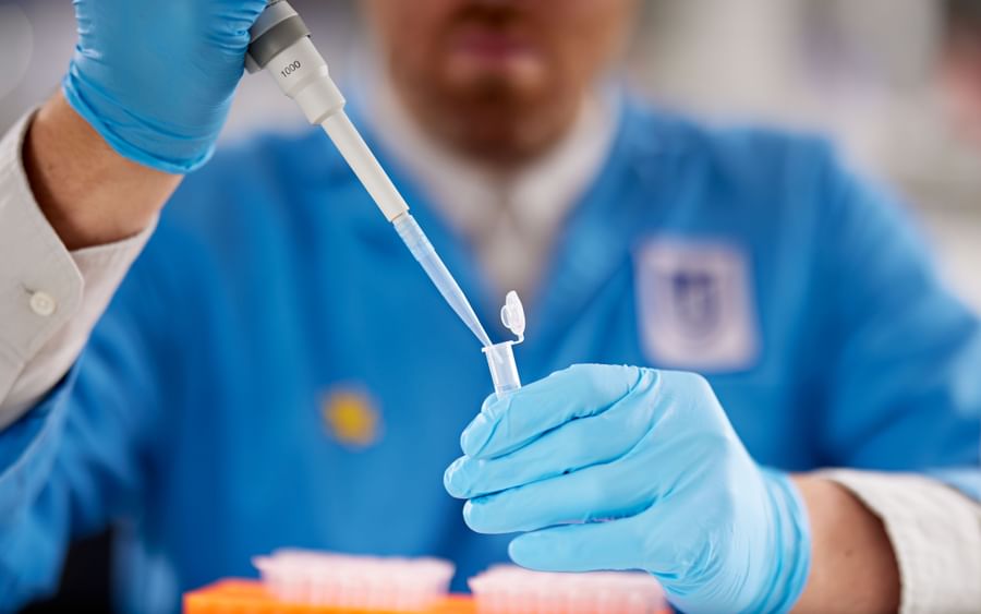 Person in blue lab coat and gloves uses a pipette to transfer liquid into a small tube.