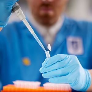 Person in blue lab coat and gloves uses a pipette to transfer liquid into a small tube.