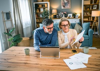 Two adults sit at a wooden table in a cosy living room, reviewing documents with a tablet.