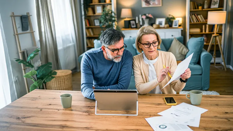 Two adults sit at a wooden table in a cosy living room, reviewing documents with a tablet.